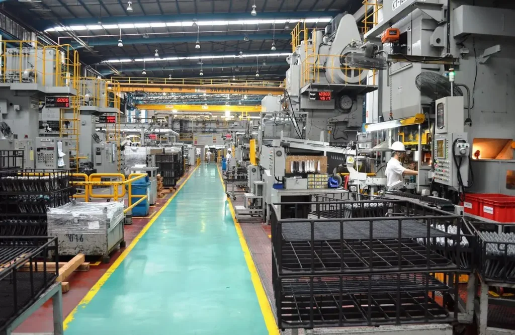 An industrial manufacturing workshop floor with heavy stamping machinery, a designated green safety walkway, and a factory worker wearing a hard hat.