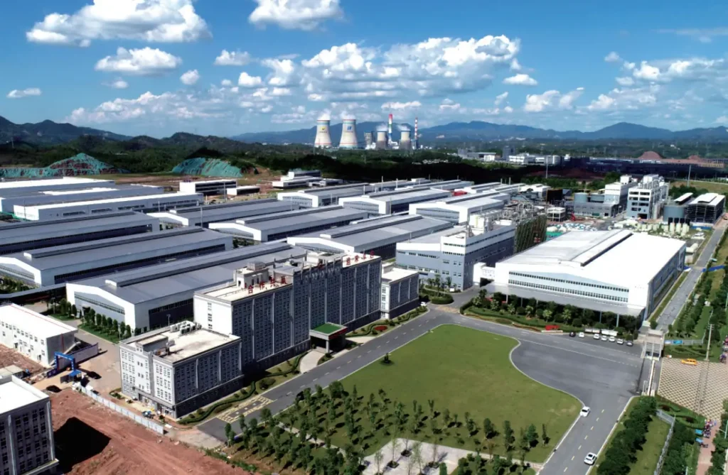An aerial drone shot of a large modern industrial park with white factory warehouses, green lawns, office buildings, and cooling towers under a blue sky.