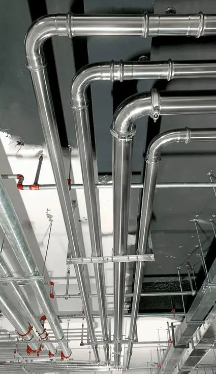 A bottom-up view of parallel stainless steel and galvanized utility pipes installed on a dark ceiling grid inside a commercial facility.