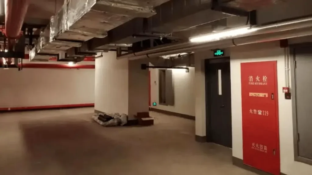 An underground building basement corridor featuring a red wall-mounted fire hydrant cabinet and ceiling-mounted ventilation and smoke exhaust ducts.