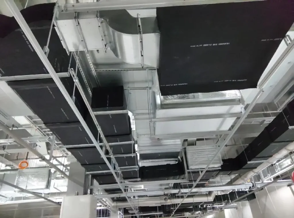 Low-angle view of industrial HVAC ventilation ductwork and insulated pipes installed on a building ceiling framework.