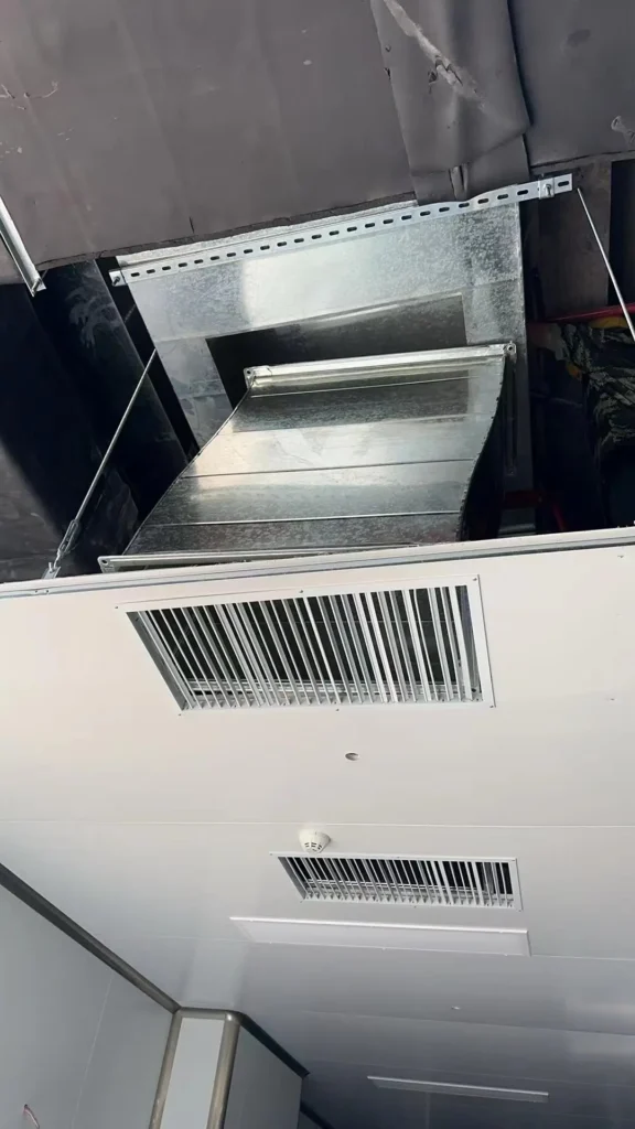 Looking up at a galvanized steel HVAC ventilation duct connecting to a linear slot diffuser through a suspended ceiling panel.
