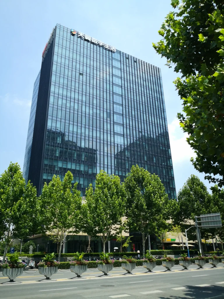 A modern multi-story commercial office building with a blue glass curtain wall facade on a sunny day with green trees in front.