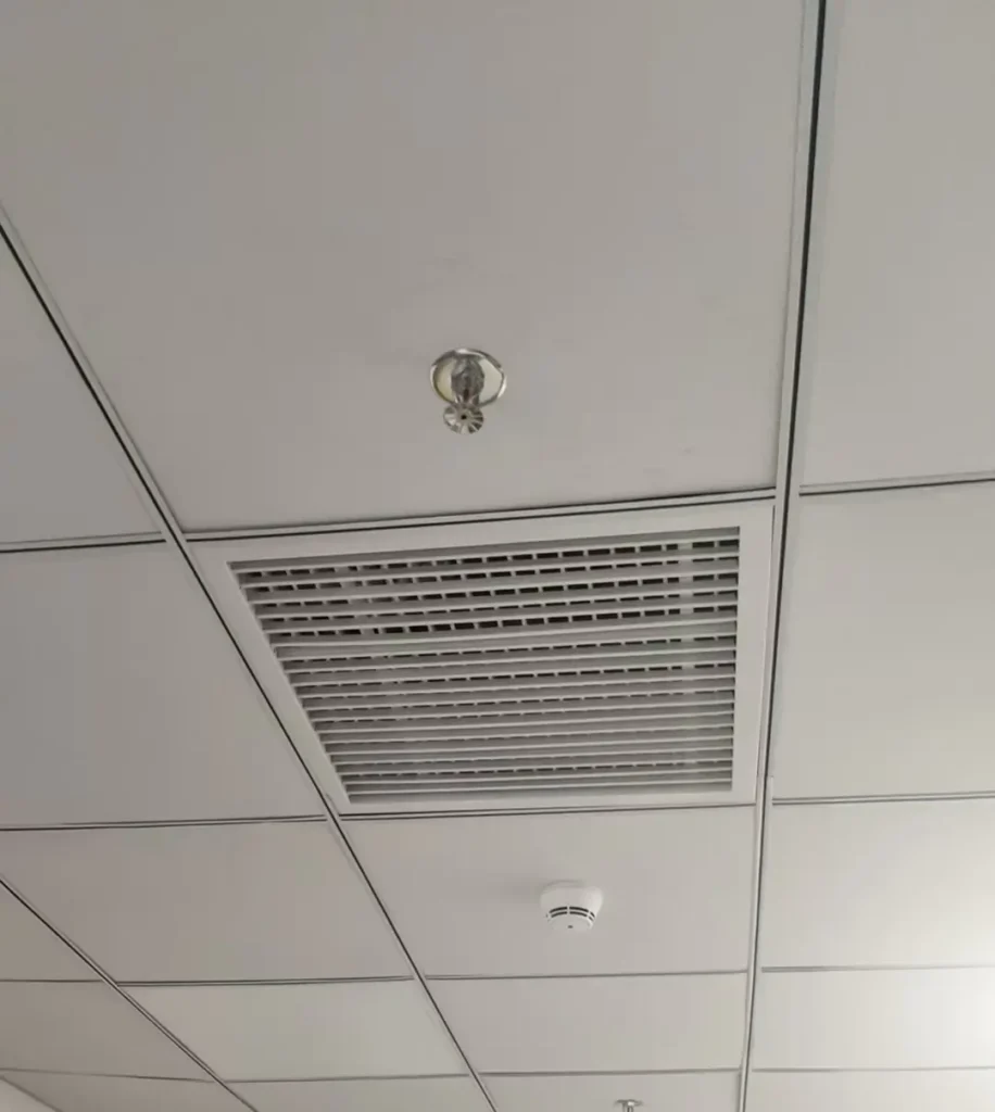 A standard office suspended ceiling panel integrated with a square HVAC air return grille, a fire sprinkler head, and a smoke detector.