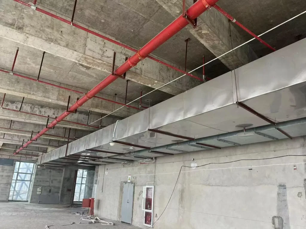 Large rectangular galvanized steel ventilation ducts and red fire sprinkler pipes hanging from a concrete ceiling during building construction.
