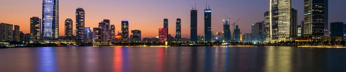 A wide panoramic night view of a modern high-rise building skyline illuminated along the waterfront, showcasing urban skyscrapers.